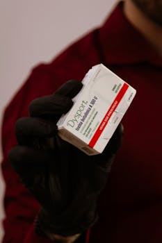 A person in red shirt holds a Dysport botulinum toxin box gloved hand.